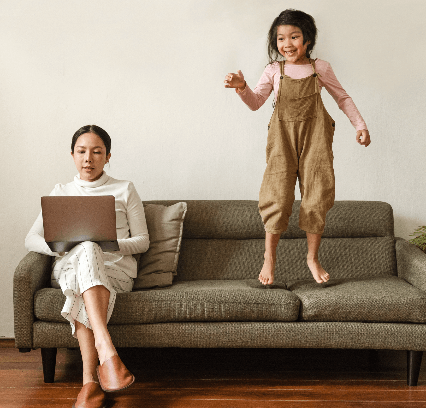 Mother on a couch with device, child jumping on the couch